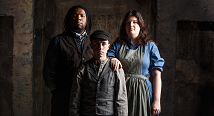 A pioneer family of three in worn 1870s clothes stands before a plaster wall in Pittsburgh Opera's Proving Up