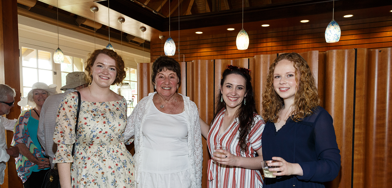 Four women pose with drinks beneath blue glass pendant lights at a FRIENDS of Pittsburgh Opera Resident Artist welcome party