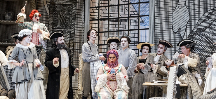 A woman with pink hair and a pink beard sits in a chair holding a jar while people in black and white clothes surround her and look at her