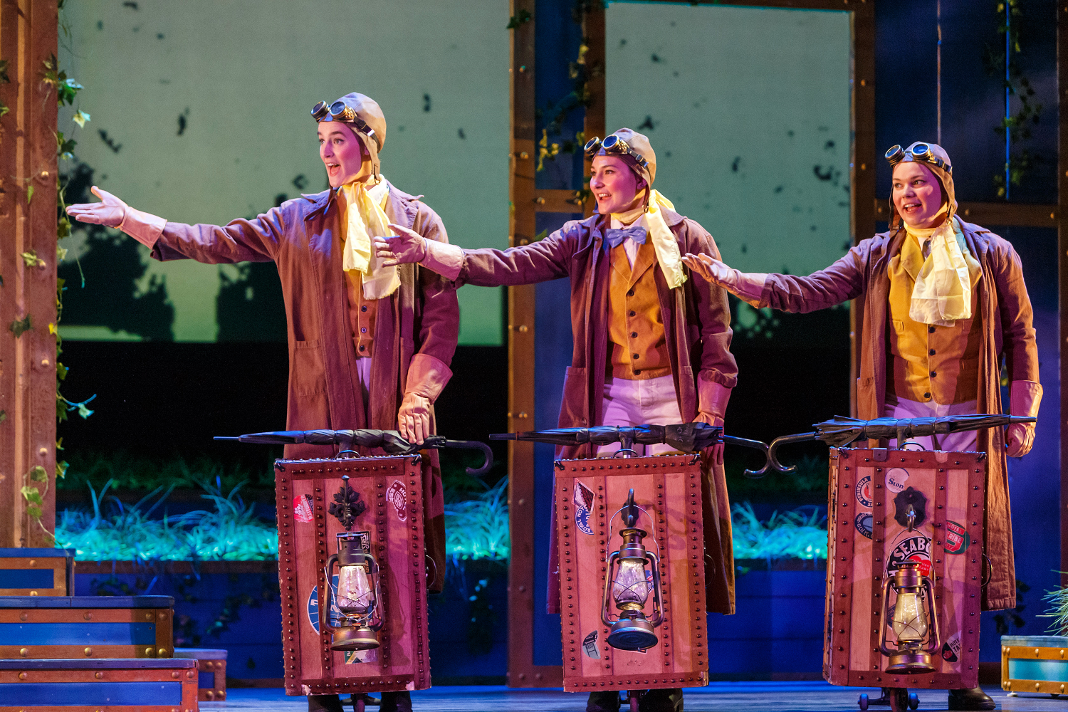 Three performers in aviator goggles and brown coats gesture from behind steamer trunks hung with lanterns