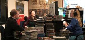 Three people talk around microphones and stacks of CDs in the WQED-FM studio