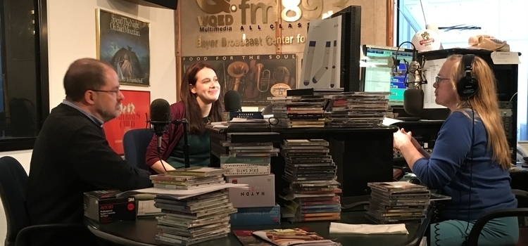 Three people talk around microphones and stacks of CDs in the WQED-FM studio