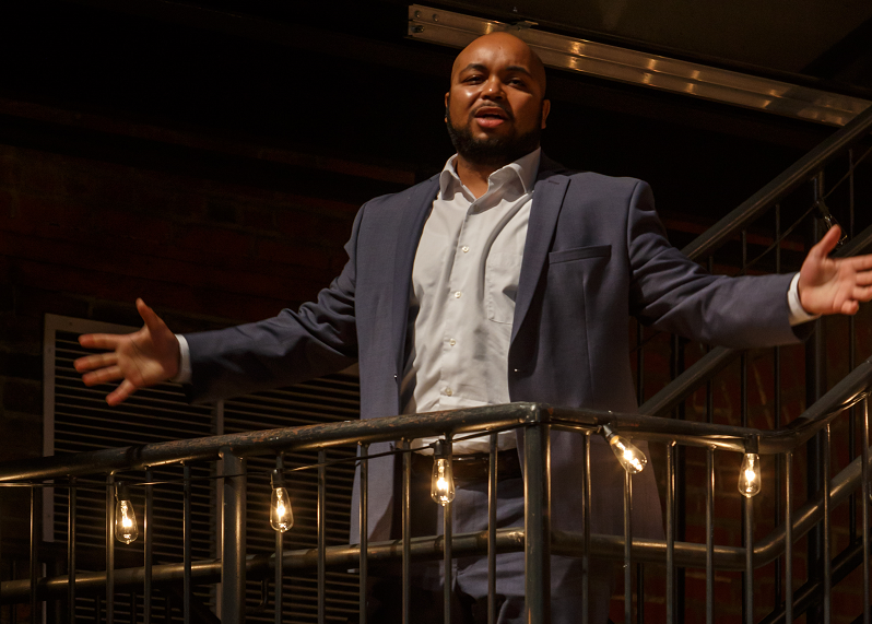 Yazid Gray in a navy suit sings with arms spread from a balcony strung with Edison bulbs at a Brown Bag concert