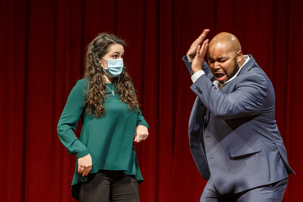 A masked soprano in teal watches Yazid Gray clap a hand to his head in a comic scene at a Brown Bag concert
