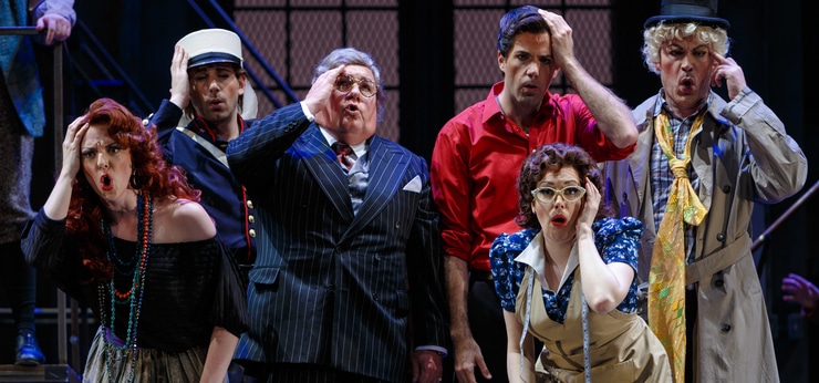 Four men and two women stand in a group and hold their hand to their face while singing. One man is dressed in a suit, another in a military uniform, another in peacoat and hat, and one in a red shirt. One of the woman iswearing a dress with a tape measure around her neck and the other wears an off-the-shoulder blouse with necklaces