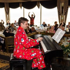 A pianist in a Christmas-print suit plays a keyboard as guests raise their arms at a FRIENDS holiday party