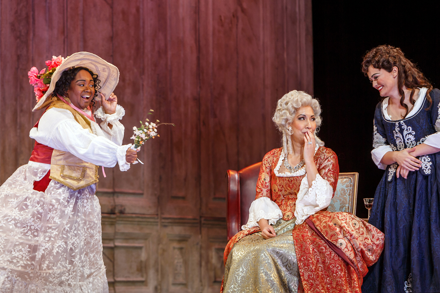 Three women in 18th-century gowns, one in a flowered bonnet waving a sprig of blossoms, on a wood-panelled set