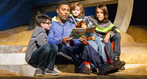A soldier in a blue hoodie reads a picture book to three children on the floor in Pittsburgh Opera's The Long Walk