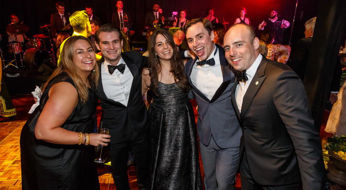 Five guests in black tie and gowns pose before the band at Maecenas, 2018