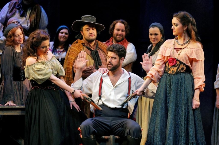 Two women draw pistols from the belt of a seated man in red suspenders as villagers raise their hands in Carmen