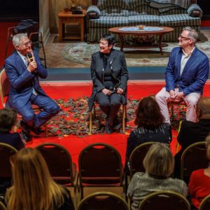 Christopher Hahn with a microphone interviews two seated artists on stage at a Meet the Artists reception