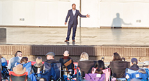 A singer in a suit performs on an outdoor amphitheater stage to a lawn-chair audience