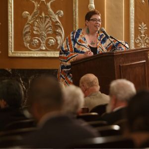 A red-haired speaker in a colorful jacket addresses an audience from a podium in the gilded Benedum Center