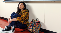 Timothi Williams as Alona sits against a locker wall beside a striped backpack in Pittsburgh Opera's Time to Act