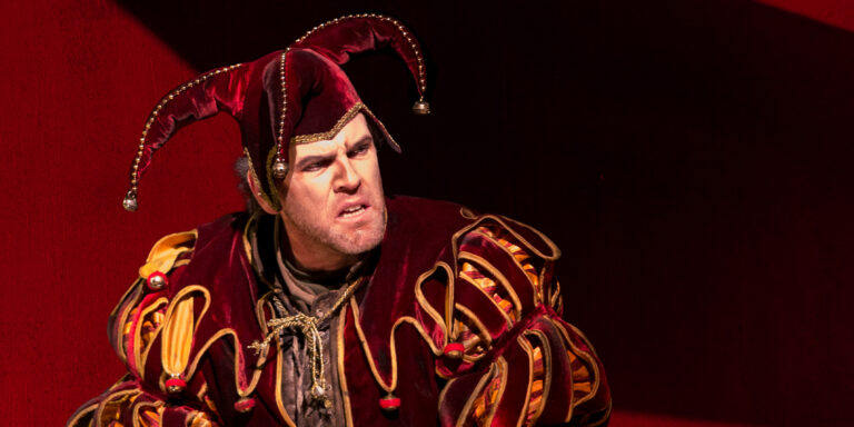 Rigoletto in a red and gold jester's costume and belled cap scowls, gripping a dagger; Rigoletto hero image
