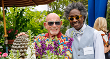 Two smiling guests in colorful attire and sunglasses pose beside purple flowers at the FRIENDS Garden Party
